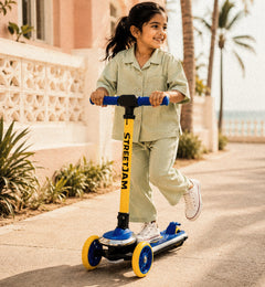 A young girl wearing a light sage green pajama set rides a blue and yellow Street Jam scooter on a sunny, sunlit sidewalk next to a pink building with palm trees.