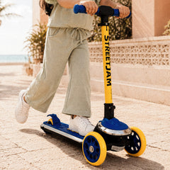 A child wearing a light sage green two-piece outfit rides a Streetjam brand scooter on a sunny outdoor patio. The scooter features a bright yellow T-bar handlebar, a blue deck, and blue wheels with yellow rims.