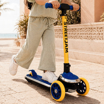 A child wearing a light sage green two-piece outfit rides a Streetjam brand scooter on a sunny outdoor patio. The scooter features a bright yellow T-bar handlebar, a blue deck, and blue wheels with yellow rims.