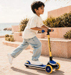 A young boy riding a yellow and blue Street Jam scooter outdoors on a sunny day near the beach, wearing a white t-shirt and light blue jeans.