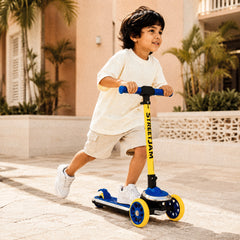 A young boy riding a yellow and blue Streetjam three-wheel scooter outdoors on a sunny day.